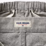 Close-up of the inside back waistband of the chef trousers showing the cream woven YOUR BRAND label with navy lettering, a red rule line and the size 32 mark, stitched into the waistband