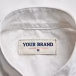 Close-up of the inside back neck of the chef uniform coat showing the cream woven YOUR BRAND label with navy lettering, a red rule line and the size M mark, stitched into the collar band