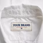 Close-up of the inside back neck of the chefs cook shirt showing the cream woven YOUR BRAND label with navy lettering, a red rule line and the size M mark, stitched into the collar band