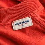 Close-up of the inside back neck of the children's wear top showing the cream woven YOUR BRAND label with navy lettering, a red rule line and the size 10Y mark, stitched into the neck binding