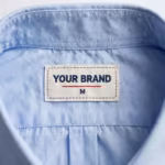 Close-up of the inside back neck of the corporate shirt showing the cream woven YOUR BRAND label with navy lettering, a red rule line and the size M mark, stitched into the collar band