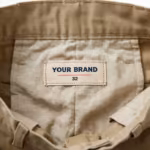 Close-up of the inside back waistband of the men's khaki cotton twill pants showing the cream woven YOUR BRAND label with navy lettering, a red rule line and the size 32 mark, stitched into the waistband