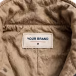 Close-up of the inside back neck of the brown duck canvas insulated coverall showing the cream woven YOUR BRAND label with navy lettering, a red rule line and the size M mark, stitched into the duck canvas neckband below the quilted collar seam