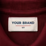 Close-up of the inside back neck of the burgundy girls' short sleeve polo shirt showing the cream woven YOUR BRAND label with navy lettering, a red rule line and the size 10Y mark, stitched into the neck band below the knitted collar