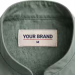 Close-up of the inside back neck of the sage green housekeeping tunic showing the cream woven YOUR BRAND label with navy lettering, a red rule line and the size M mark, stitched into the inside back neck below the stand collar seam