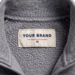 Close-up of the inside back neck of the grey marl brushed fleece jogging suit jacket showing the cream woven YOUR BRAND label with navy lettering, a red rule line and the size M mark, stitched into the brushed loopback fleece below the ribbed stand collar