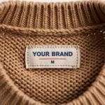 Close-up of the inside back neck of the camel knitted crew-neck sweater showing the cream woven YOUR BRAND label with navy lettering, a red rule line and the size M mark, stitched into the inside back neck just below the ribbed crew neck band