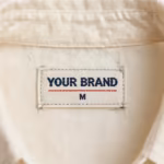 Close-up of the inside back neck of the ivory woven rayon blouse showing the cream woven YOUR BRAND label with navy lettering, a red rule line and the size M mark, stitched into the woven neck binding
