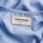 Close-up of the inside back neck of the ceil blue ladies cross over scrub top showing the cream woven YOUR BRAND label with navy lettering, a red rule line and the size M mark, stitched into the inside back neck below the neck binding