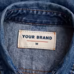 Close-up of the inside back neck of the ladies denim top showing the cream woven YOUR BRAND label with navy lettering, a red rule line and size M mark, stitched into the neck seam with copper topstitching on indigo denim