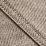 Close-up of the stone beige cotton twill weave with fine diagonal twill lines, natural fibre texture and a felled seam with two stitching rows