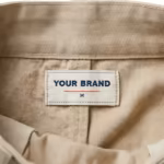Close-up of the inside back waistband of the ladies' A-line midi skirt in stone beige cotton twill showing the cream woven YOUR BRAND label with navy lettering, a red rule line and the size M mark, stitched into the waistband