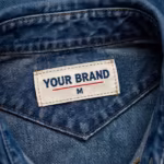 Close-up of the inside back neck of the denim shirt showing the cream woven YOUR BRAND label with navy lettering, a red rule line and the size M mark, stitched into the collar band