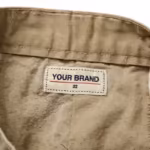 Close-up of the inside back waistband of the men's khaki casual chino shorts showing the cream woven YOUR BRAND label with navy lettering, a red rule line and the size 32 mark, stitched into the waistband