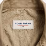 Close-up of the inside back neck of the men's khaki cotton twill chore jacket showing the cream woven YOUR BRAND label with navy lettering, a red rule line and the size M mark, stitched into the centre of the inside back neck below the collar on the reverse side of the khaki twill