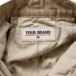 Close-up of the inside back waistband of the men's khaki heavy twill work cargo shorts showing the cream woven YOUR BRAND label with navy lettering, a red rule line and the size 32 mark, stitched into the waistband