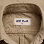 Close-up of the inside back neck of the men's khaki cotton twill work shirt showing the cream woven YOUR BRAND label with navy lettering, a red rule line and the size M mark, stitched into the inside collar band