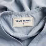 Close-up of the inside back neck of the ceil blue men's zip pocket scrub top showing the cream woven YOUR BRAND label with navy lettering, a red rule line and the size M mark, stitched into the inside back neckband