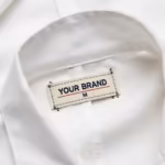 Close-up of the inside back neck of the men's white zipper smock lab coat showing the cream woven YOUR BRAND label with navy lettering, a red rule line and the size M mark, stitched into the inside back neck below the stand collar