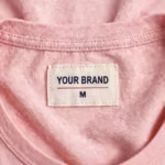 Close-up of the inside back neck of the ladies soft pink night t-shirt showing the cream woven YOUR BRAND label with navy lettering, a red rule line and the size M mark, stitched into the bound crew neck binding
