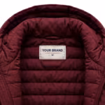 Close-up of the inside back neck of the burgundy quilted padded jacket showing the cream woven YOUR BRAND label with navy lettering, a red rule line and the size M mark, stitched into the quilted burgundy lining below the collar