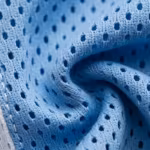 Close-up of the sky blue polyester eyelet mesh of the sportswear range, showing the perforated mesh holes, knit loops and yarn fibres across a soft fold, with a white mesh panel along one edge
