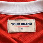 Close-up of the inside back neck of the red team jersey in polyester eyelet mesh showing the cream woven YOUR BRAND label with navy lettering, a red rule line and the size M mark, stitched into the inside back neck just below the white ribbed collar tape