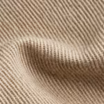 Close-up of the stone beige cotton twill weave of the trousers range, showing the diagonal ribs, yarn structure and fibre fly across a soft fold