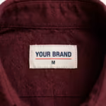 Close-up of the inside back neck of the burgundy poly-cotton twill staff uniform shirt showing the cream woven YOUR BRAND label with navy lettering, a red rule line and the size M mark, stitched into the inside back neck below the collar seam
