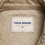 Close-up of the inside back neck of the men's khaki zip front cotton coverall showing the cream woven YOUR BRAND label with navy lettering, a red rule line and the size M mark, stitched into the inside back neck below the stand collar