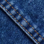Mid-indigo denim fabric close-up showing the diagonal twill weave, yarns and a felled seam