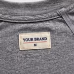 Close-up of the inside back neck of the corporate t-shirt showing the cream woven YOUR BRAND label with navy lettering, a red rule line and the size M mark, stitched into the neck binding