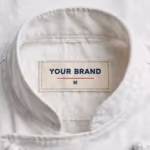 Close-up of the inside back neck of the white executive chef coat, showing the cream woven YOUR BRAND label with navy lettering, a red rule line and the size M mark, stitched into the collar band