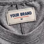 Close-up of the inside back neck of the grey marl gym training t-shirt showing the cream woven YOUR BRAND label with navy lettering, a red rule line and the size M mark, stitched into the ribbed crew neck band