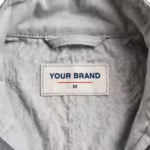 Close-up of the inside back neck of the men's stone grey industrial coverall showing the cream woven YOUR BRAND label with navy lettering, a red rule line and the size M mark, stitched into the neck seam below the stand collar