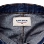 Close-up of the inside back waistband of the men's mid indigo jeans pants showing the cream woven YOUR BRAND label with navy lettering, a red rule line and the size 32 mark, stitched into the waistband