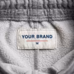 Close-up of the inside back waistband of the grey marl jogging pants showing the cream woven YOUR BRAND label with navy lettering, a red rule line and the size M mark, stitched into the waistband