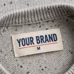 Close-up of the inside back neck of the men's light grey marl knitted crew neck jumper showing the cream woven YOUR BRAND label with navy lettering, a red rule line and the size M mark, stitched into the reverse stocking stitch below the ribbed crew neck band