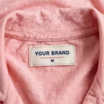 Close-up of the inside back neck of the soft pink pyjama top showing the cream woven YOUR BRAND label with navy lettering, a red rule line and the size M mark, stitched into the inside back neck below the collar band