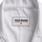 Close-up of the inside back neck of the white lab coat showing the cream woven YOUR BRAND label with navy lettering, a red rule line and the size M mark, stitched into the neckband below the collar