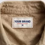 Close-up of the inside back neck of the khaki canvas work shirt showing the cream woven YOUR BRAND label with navy lettering, a red rule line and the size M mark, stitched below the collar band