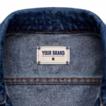 Close-up of the inside back neck of the denim jacket showing the cream woven YOUR BRAND label with navy lettering, a red rule line and the size M mark, stitched into the collar band