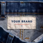 Close-up of the inside back waistband of the indigo denim trousers, showing the cream woven YOUR BRAND label with navy lettering, a red rule line and the size 32 mark, stitched into the waistband
