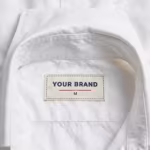 Close-up of the inside back neck of the white dress shirt showing the cream woven YOUR BRAND label with navy lettering, a red rule line and size M mark, stitched in below the collar band