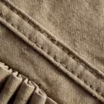 Close-up of the khaki cotton canvas weave of the multi pocket vest, showing the yarn structure and fibre fly, a felled seam with double stitching and a bellows pleat fold