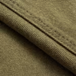 Close-up of the olive cotton twill weave of the parka shell, showing individual yarns, natural fibres, a soft fold and a stitched seam