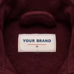 Close-up of the inside back neck of the men's burgundy full-zip polar fleece jacket showing the cream woven YOUR BRAND label with navy lettering, a red rule line and the size M mark, stitched into the inside back neck just below the stand collar on the reverse side of the burgundy polar fleece