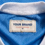 Close-up of the inside back neck of the men's sky blue soccer jersey showing the cream woven YOUR BRAND label with navy lettering, a red rule line and the size M mark, stitched into the sky blue eyelet mesh just below the white ribbed crew collar