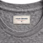 Close-up of the inside back neck of the grey marl classic crew neck t-shirt showing the cream woven YOUR BRAND label with navy lettering, a red rule line and the size M mark, stitched into the inside back neck below the ribbed crew collar