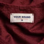 Close-up of the inside of the burgundy work tabard with pockets below the neck edge showing the cream woven YOUR BRAND label with navy lettering, a red rule line and the size M mark, stitched into the tabard's own fabric